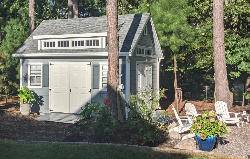A small shed with a patio featuring two chairs, surrounded by greenery and a clear blue sky.