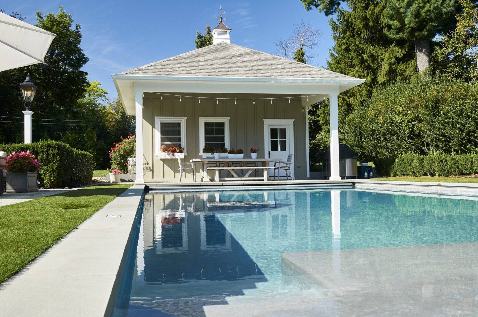 A pool house featuring a patio with a table, surrounded by greenery and a clear blue sky.
