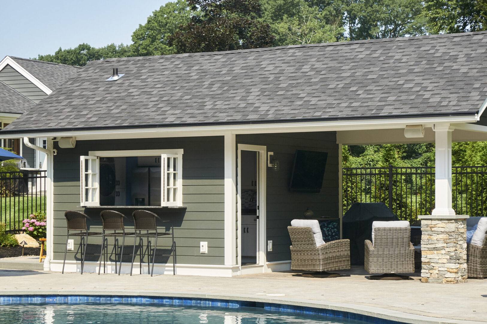 A pool house featuring a spacious patio and a grill, surrounded by greenery and outdoor furniture.